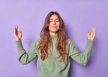 woman holds hands zen om gesture keeps eyes closed meditates indoor dressed casually keeps emotions control peace patience breathes air freely isolated purple wall
