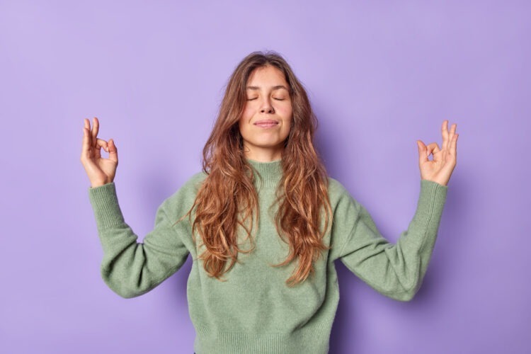 woman holds hands zen om gesture keeps eyes closed meditates indoor dressed casually keeps emotions control peace patience breathes air freely isolated purple wall