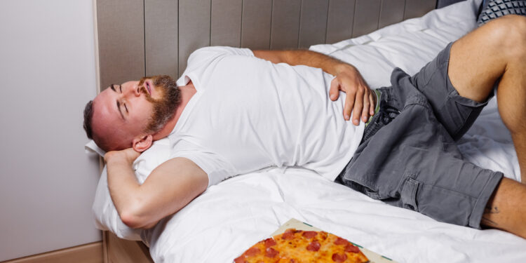 Caucasian bearded man on a bed after overindulging in fast food pizza, highlighting the link between poor Gut Health and Sleep dietary choices and sleep quality