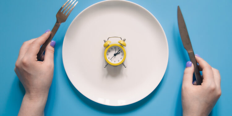 Hands holding knife and fork above alarm clock on a plate on blue background. Intermittent fasting, Ketogenic dieting, weight loss, meal plan, and healthy food concept.